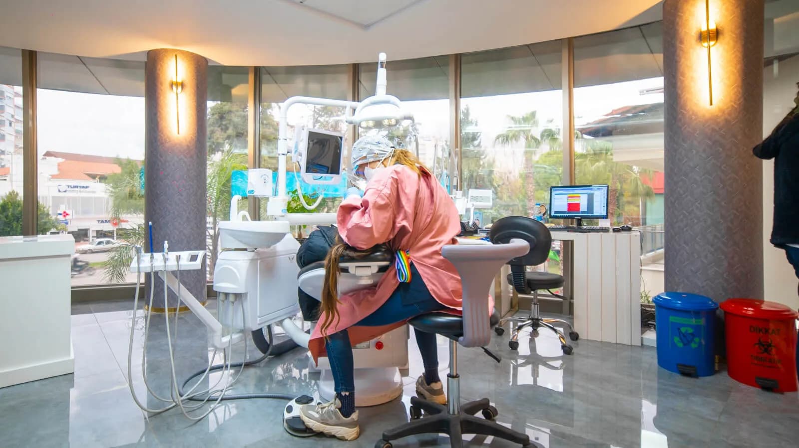 Eine Behandlerin mit Mundschutz bei Ballıpınar Dental behandelt eine Patientin in einem hellen Behandlungsraum, beide von hinten zu sehen