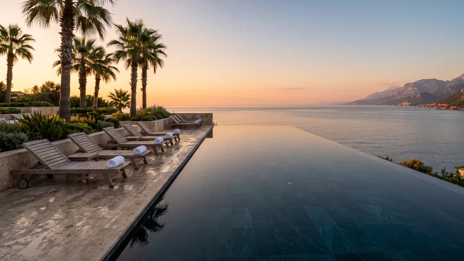 Eine Hotel-Poolterrasse im Abendlicht, leere Liegen unter Palmen, dahinter das Mittelmeer